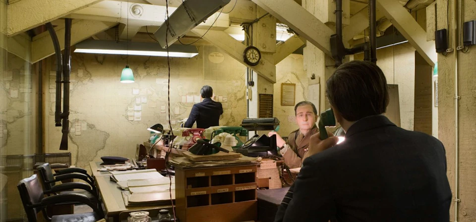 Two men in a dimly lit, cluttered office with industrial decor; one man in military attire, the other in a suit, using a phone.