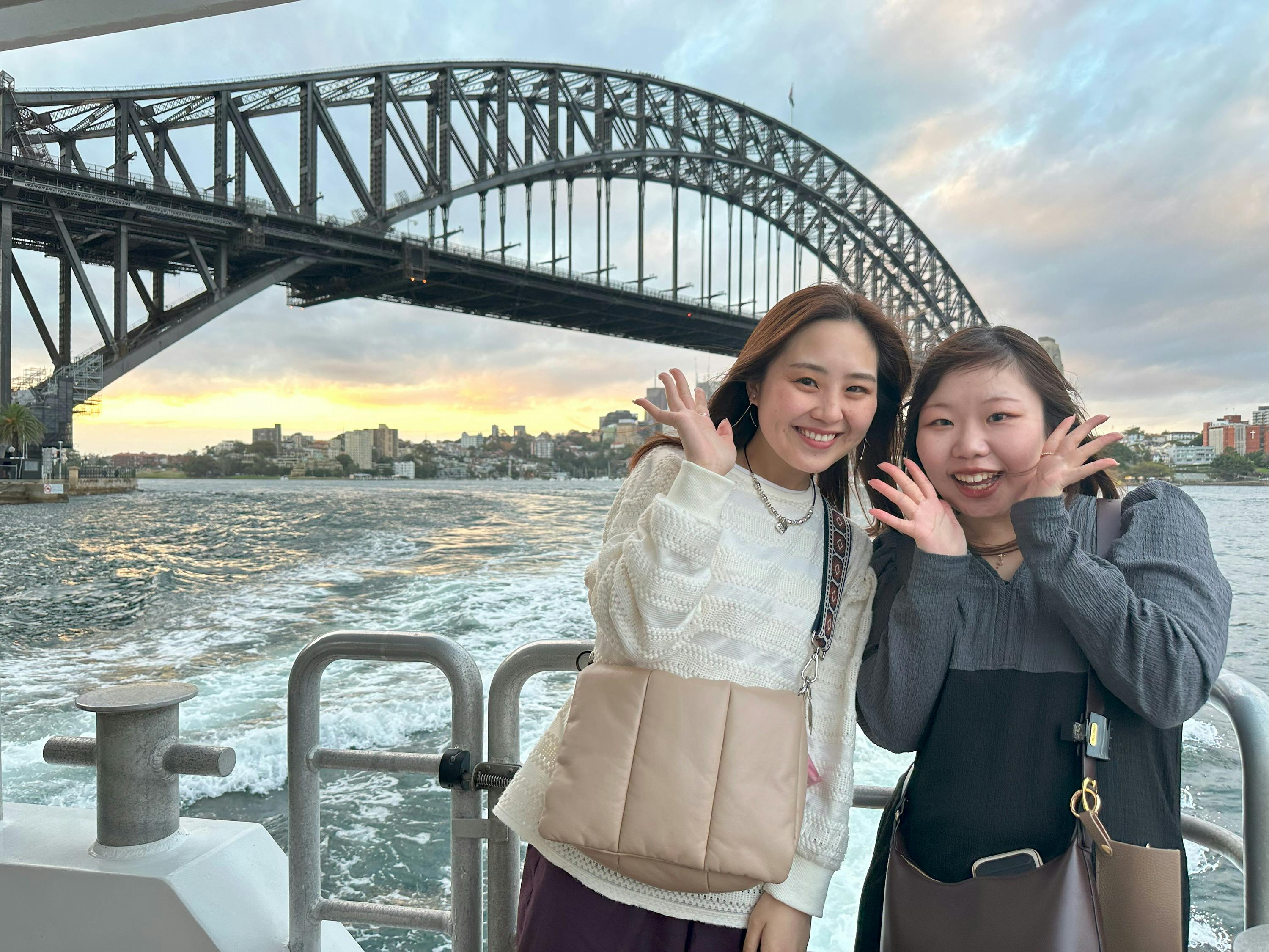 Two women smiling and waving on a boat with the Sydney Harbour Bridge and cityscape in the background during sunset.