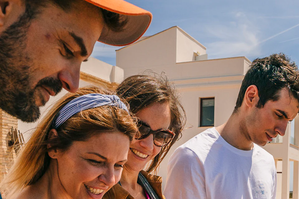 Four people read an informational sign outdoors near white buildings on a sunny day.