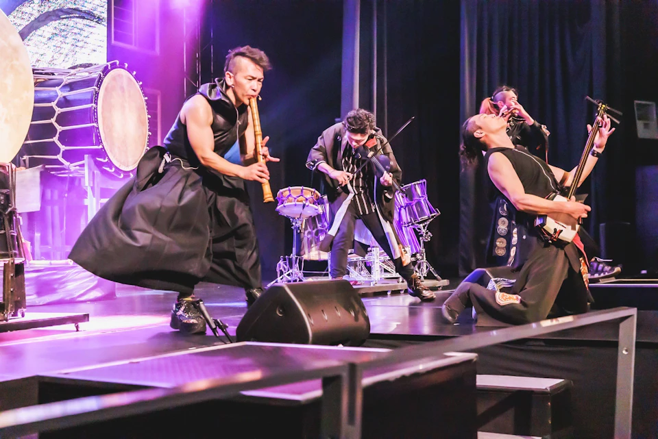 Two performers on stage, one playing a bamboo flute and the other a violin, with drums and stage lighting in the background.