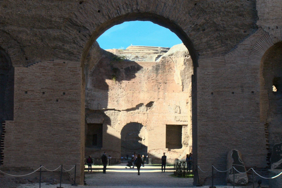 An ancient brick archway revealing ruins with more arches, people walking, and a clear blue sky.