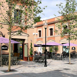 Utomhuscafé med lila paraplyer, träbord och stolar på ett asfalterat torg. Solbrända byggnader med stora fönster och träd omger området.