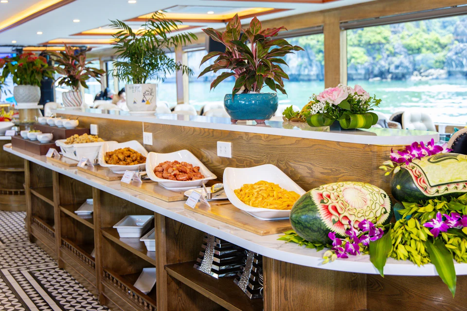 A buffet table with various dishes, decorative plants, and a carved watermelon, set in a well-lit dining area with scenic window views.