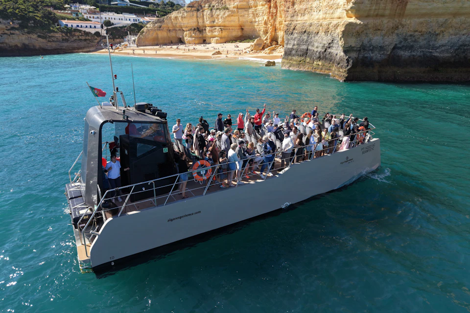 A boat with people on board sailing near a rocky coastline and a sandy beach under a clear sky.