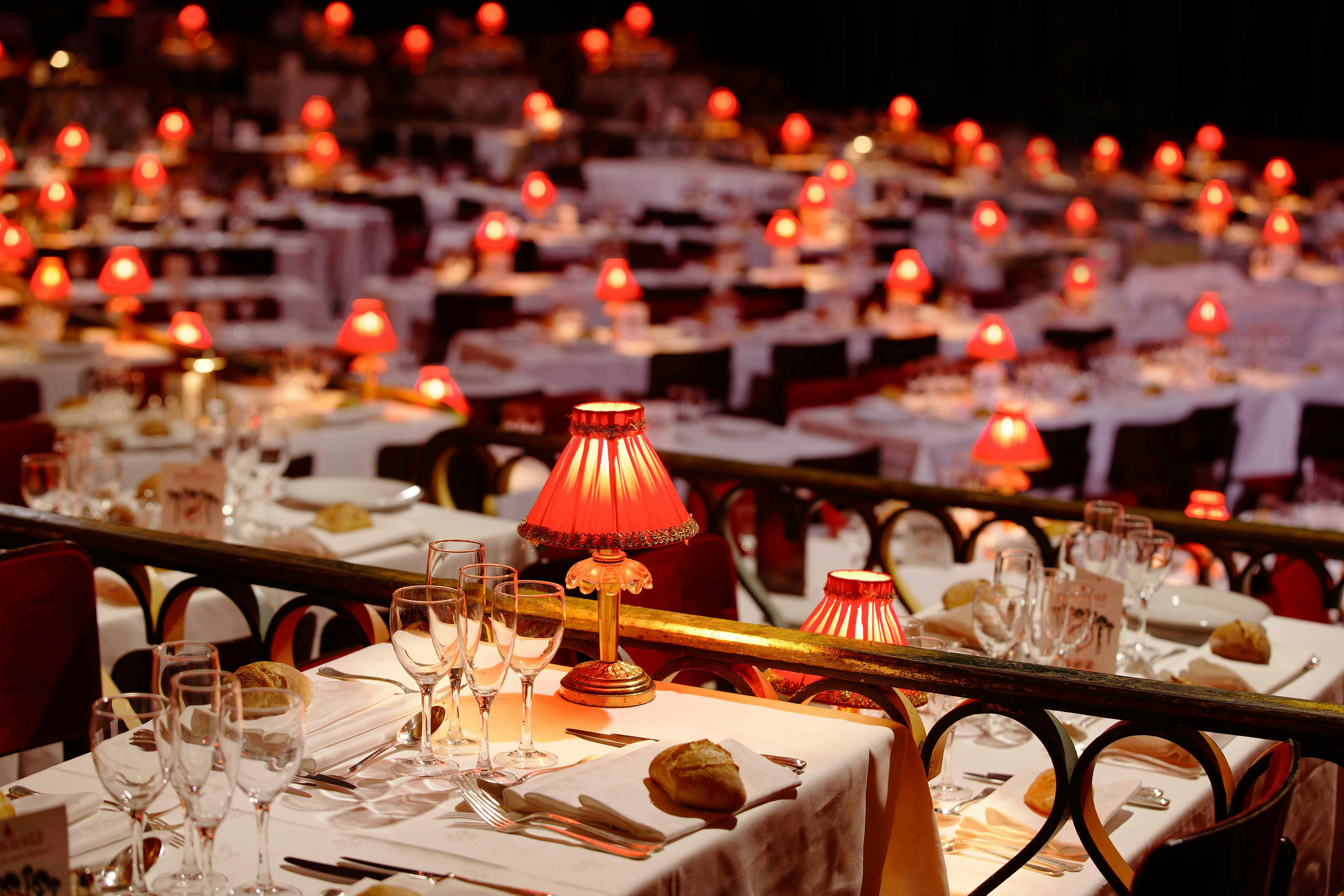 A dimly lit restaurant or banquet hall set with white tablecloths, red-shaded lamps, glassware, and bread rolls on the tables.