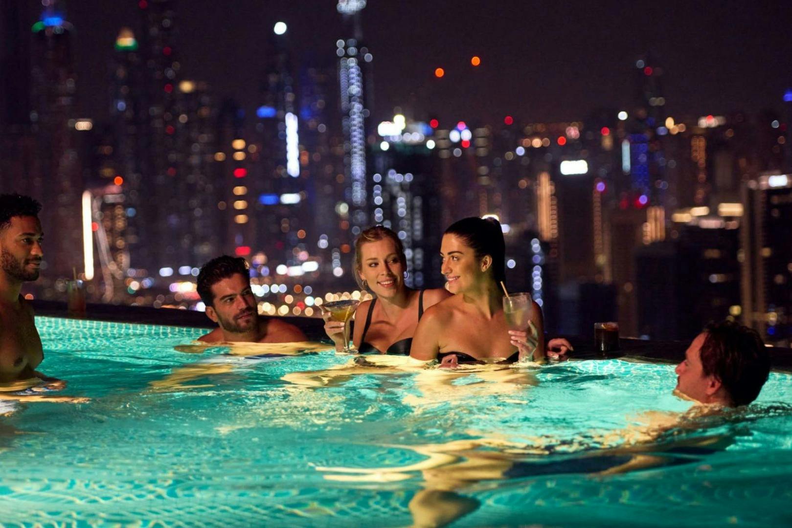 Four people in a rooftop infinity pool at night, holding drinks and chatting, with a brightly lit city skyline in the background.