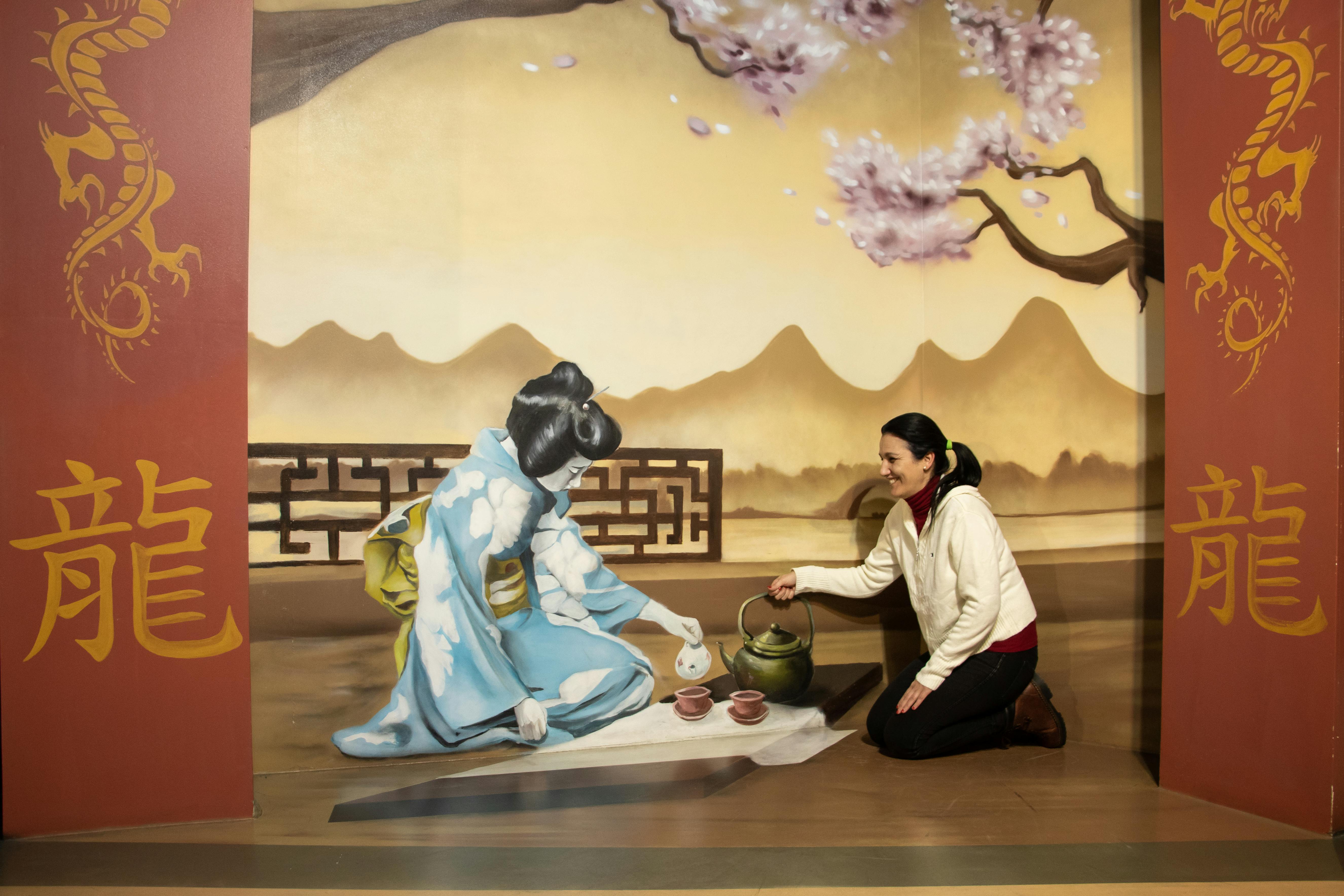 Une femme interagit avec une peinture murale représentant une scène de thé traditionnelle japonaise sur fond de montagnes et de cerisiers en fleurs.