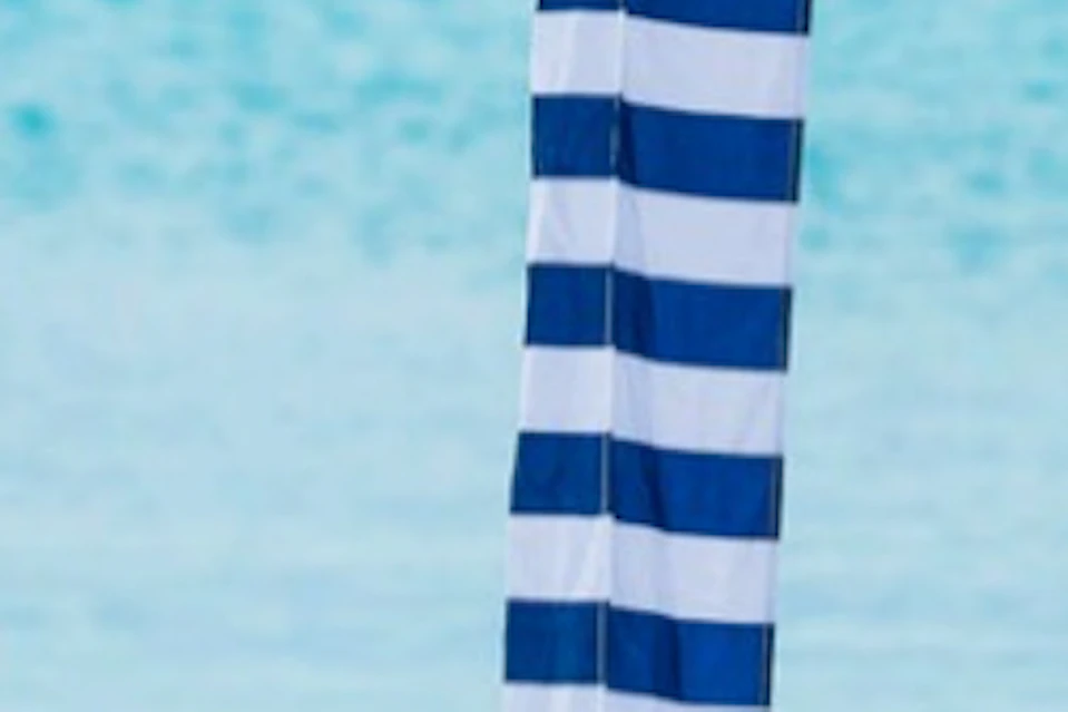 Two people sitting on a beach under a blue and white striped canopy, with clear turquoise water and boats in the background.