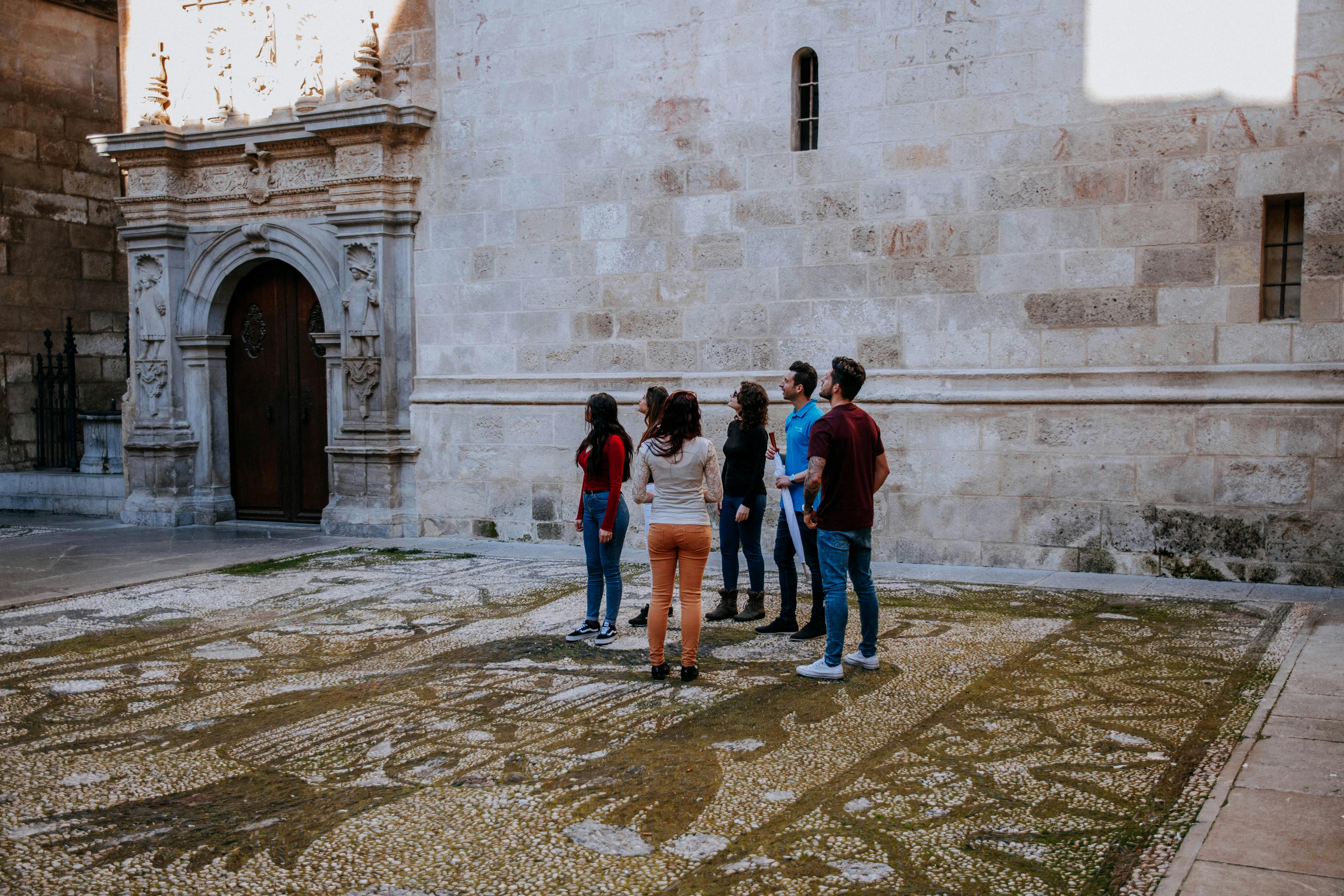 Skupina šesti lidí stojí před historickou kamennou budovou s ozdobnými dřevěnými dveřmi a dívá se vzhůru.