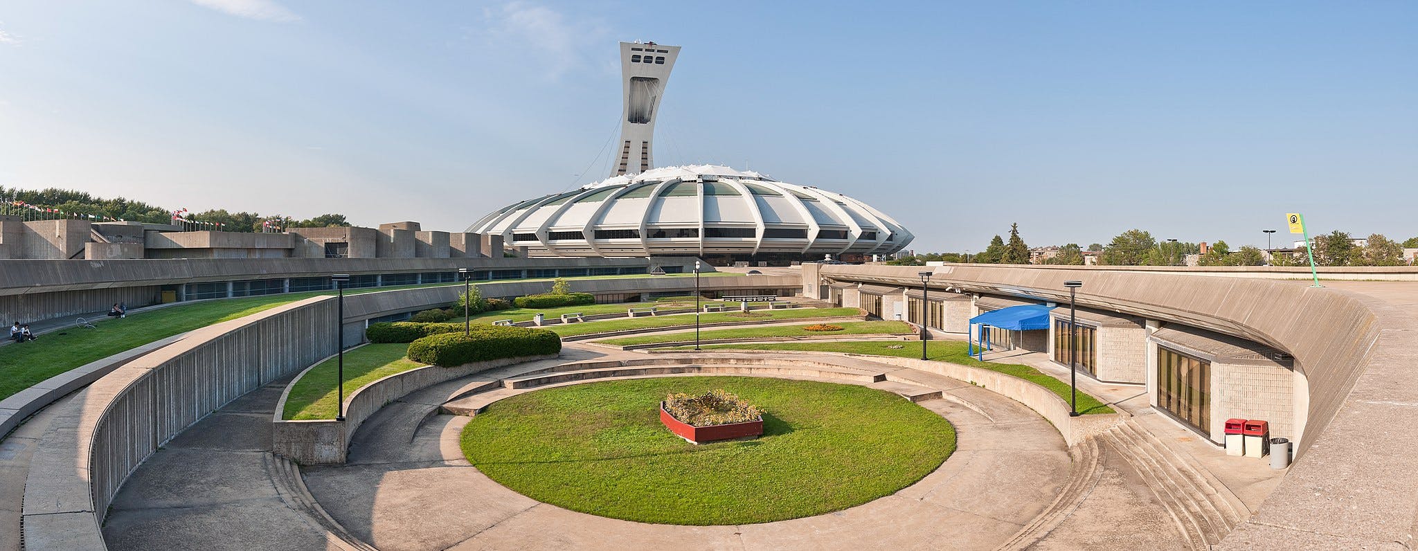 Circular concrete and grassy tiers in front of a large, dome-shaped building with an inclined tower in a sunny, outdoor setting.
