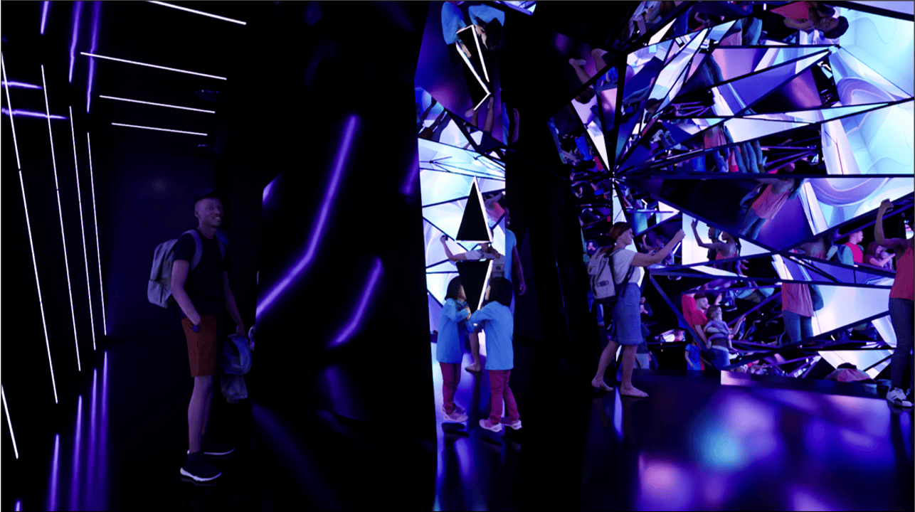 A mirrored room with purple lighting, children and adults interacting with reflective surfaces and enjoying the space.