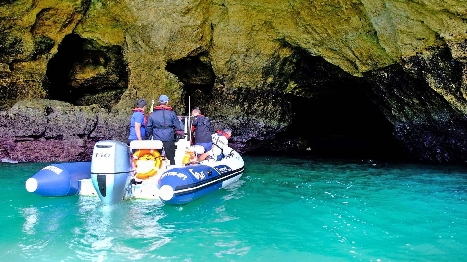 Three people in a small boat approach the entrance of a rocky cave on a turquoise water body.
