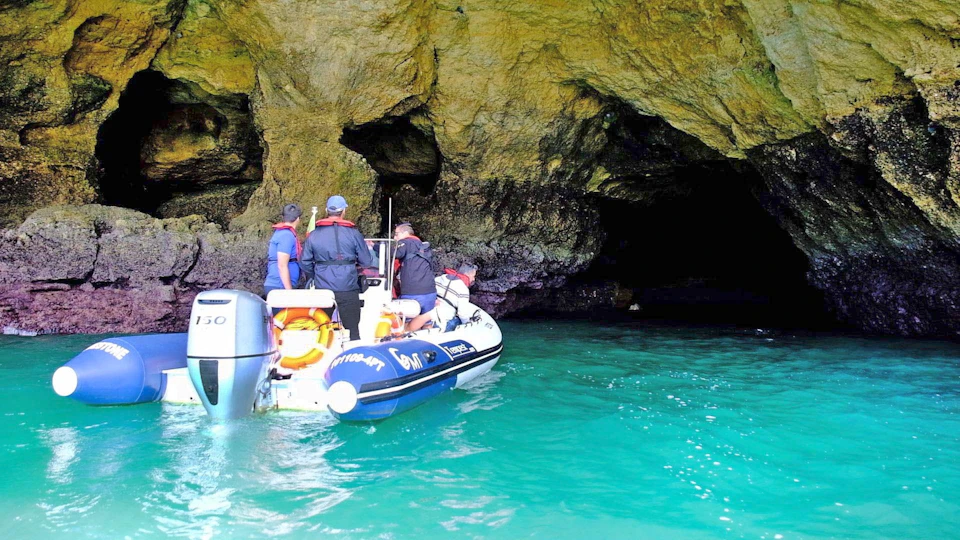 Three people in a small boat approach the entrance of a rocky cave on a turquoise water body.