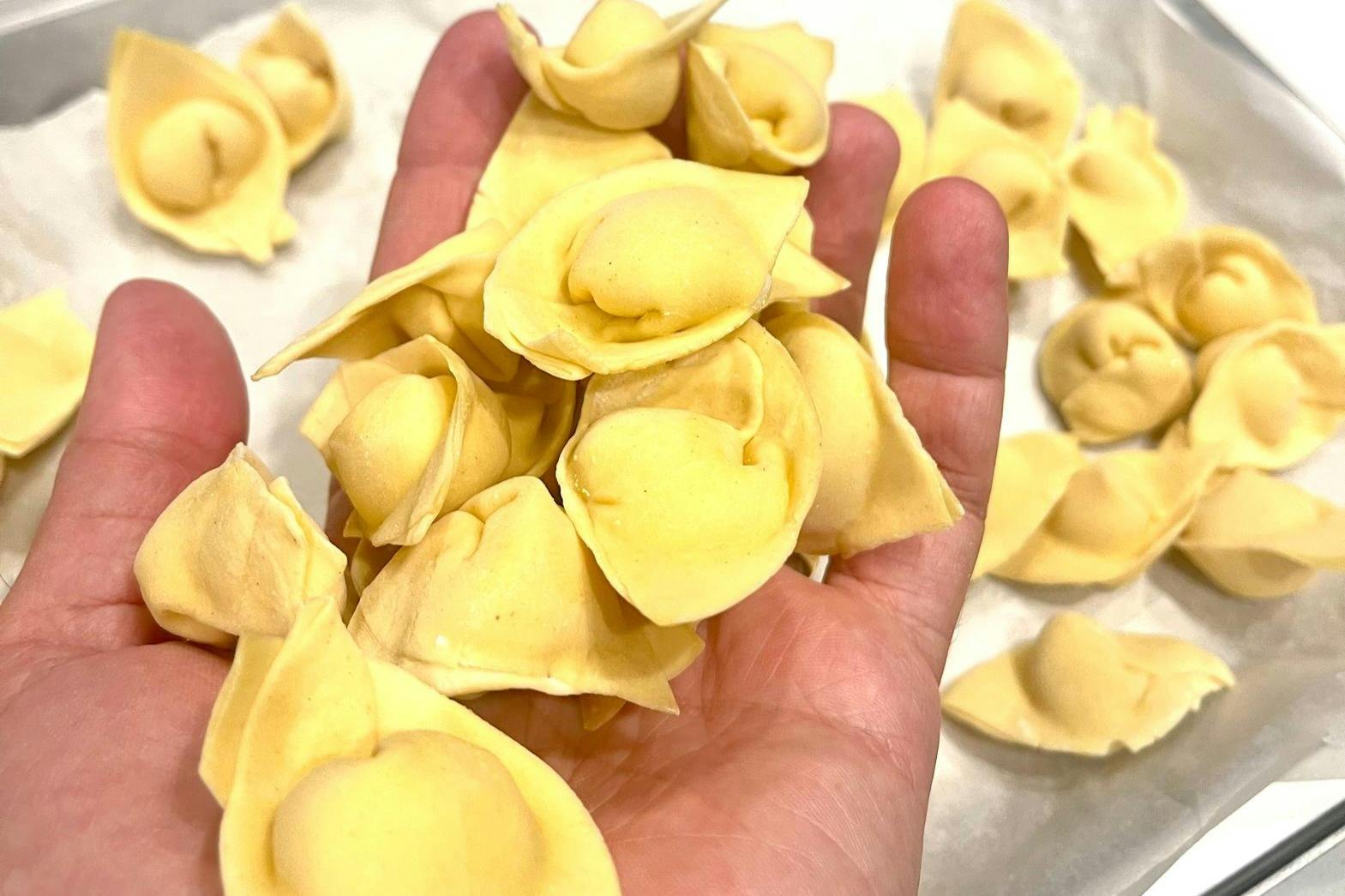 A hand holding a bunch of uncooked tortellini pasta, with more tortellini scattered on a white surface in the background.