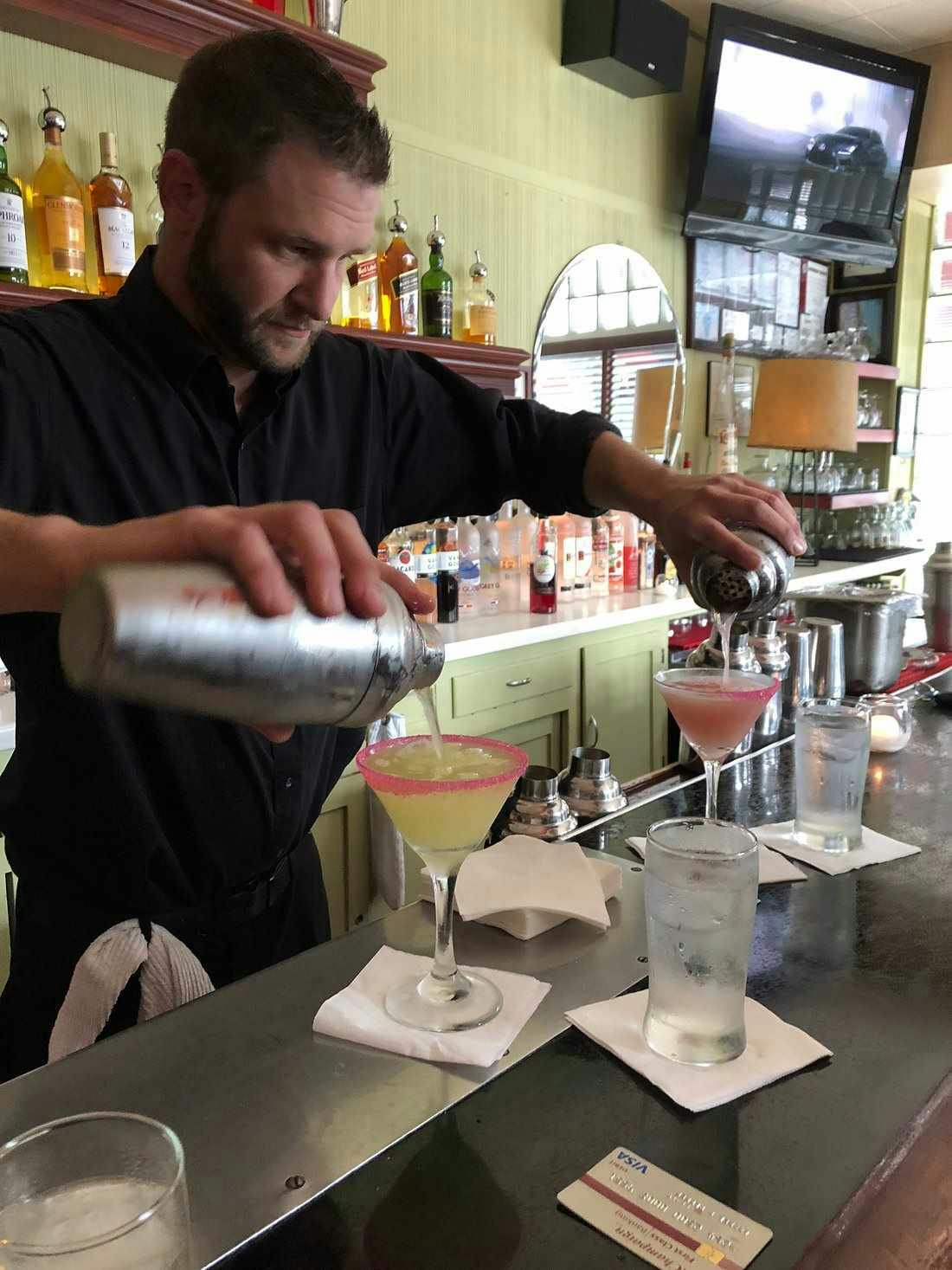 Bartender pouring two cocktails simultaneously from shakers at a bar, with bottles and a TV in the background.