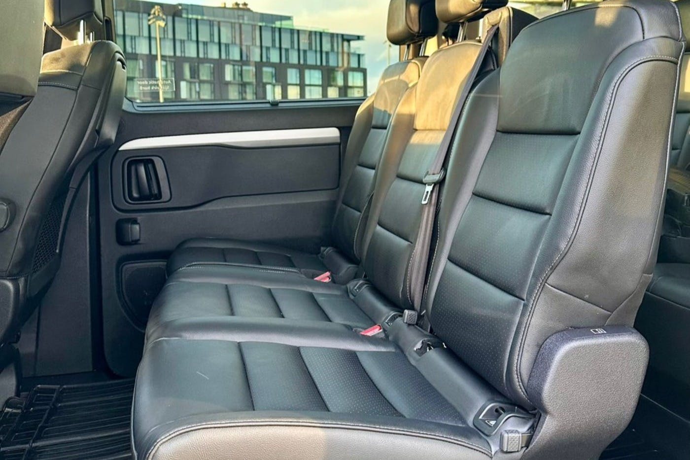 Backseat view of a vehicle with three black leather seats. Sunlight filters in, and a modern building is visible through the window.