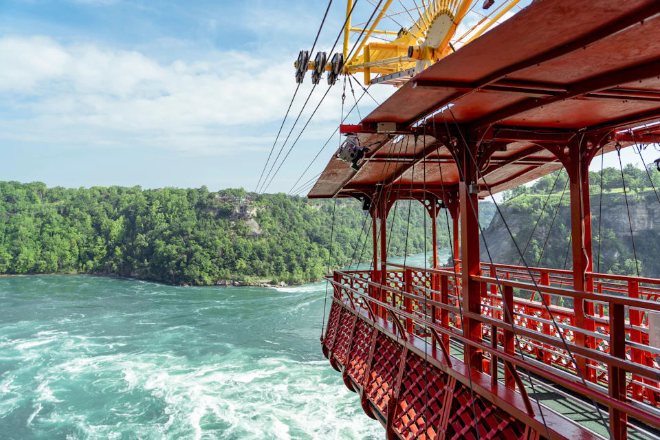 A red cable car suspended over a river with rapids, surrounded by lush green forested cliffs under a partly cloudy sky.