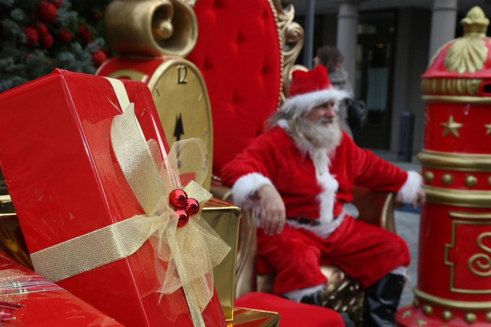 A person dressed as Santa Claus sits on a large, ornate red and gold chair, surrounded by festive holiday decorations.