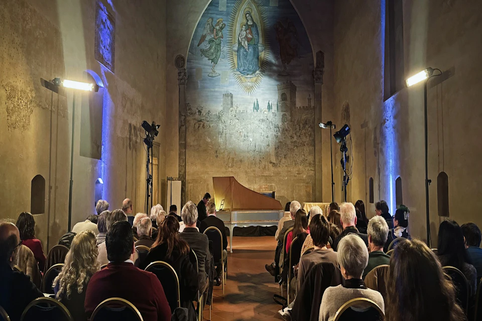 Audience seated in a hall facing a harpsichord and a painted mural on the wall, lit by soft lighting.