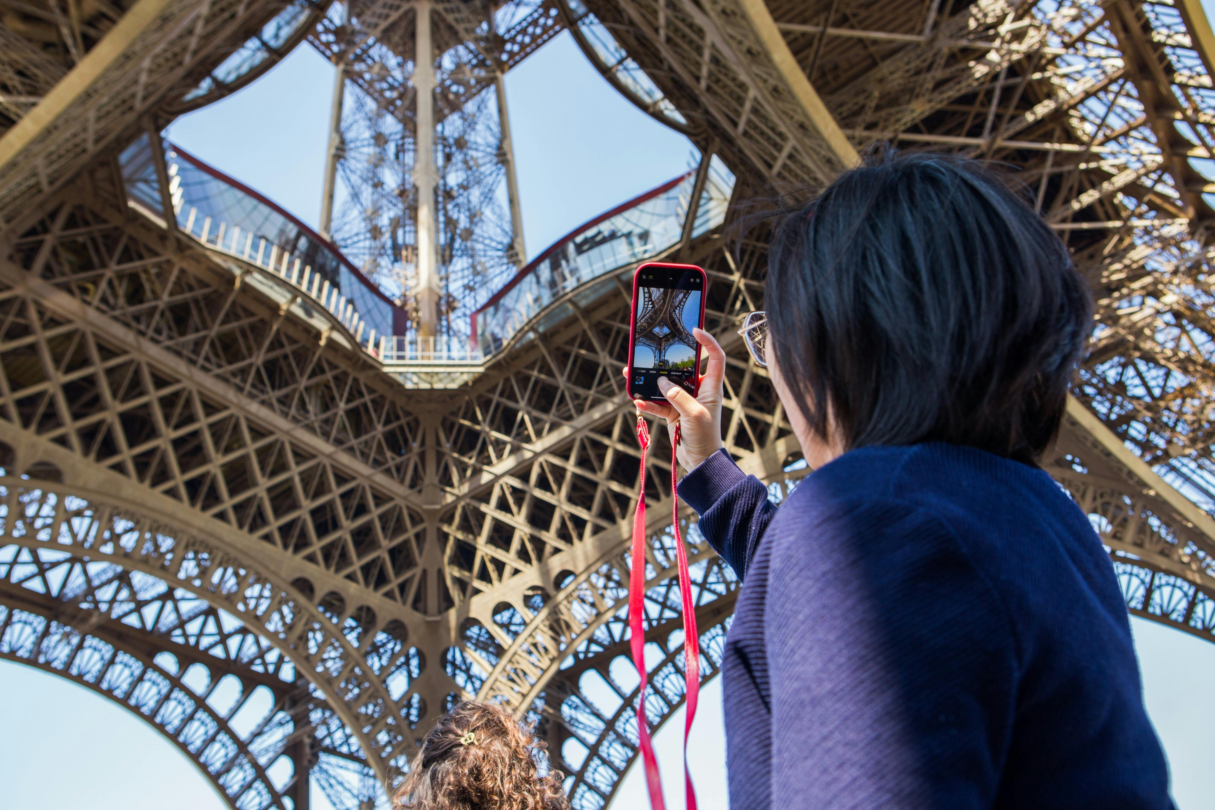 A person holding a smartphone takes a photo of the intricate lattice structure of the Eiffel Tower from below.