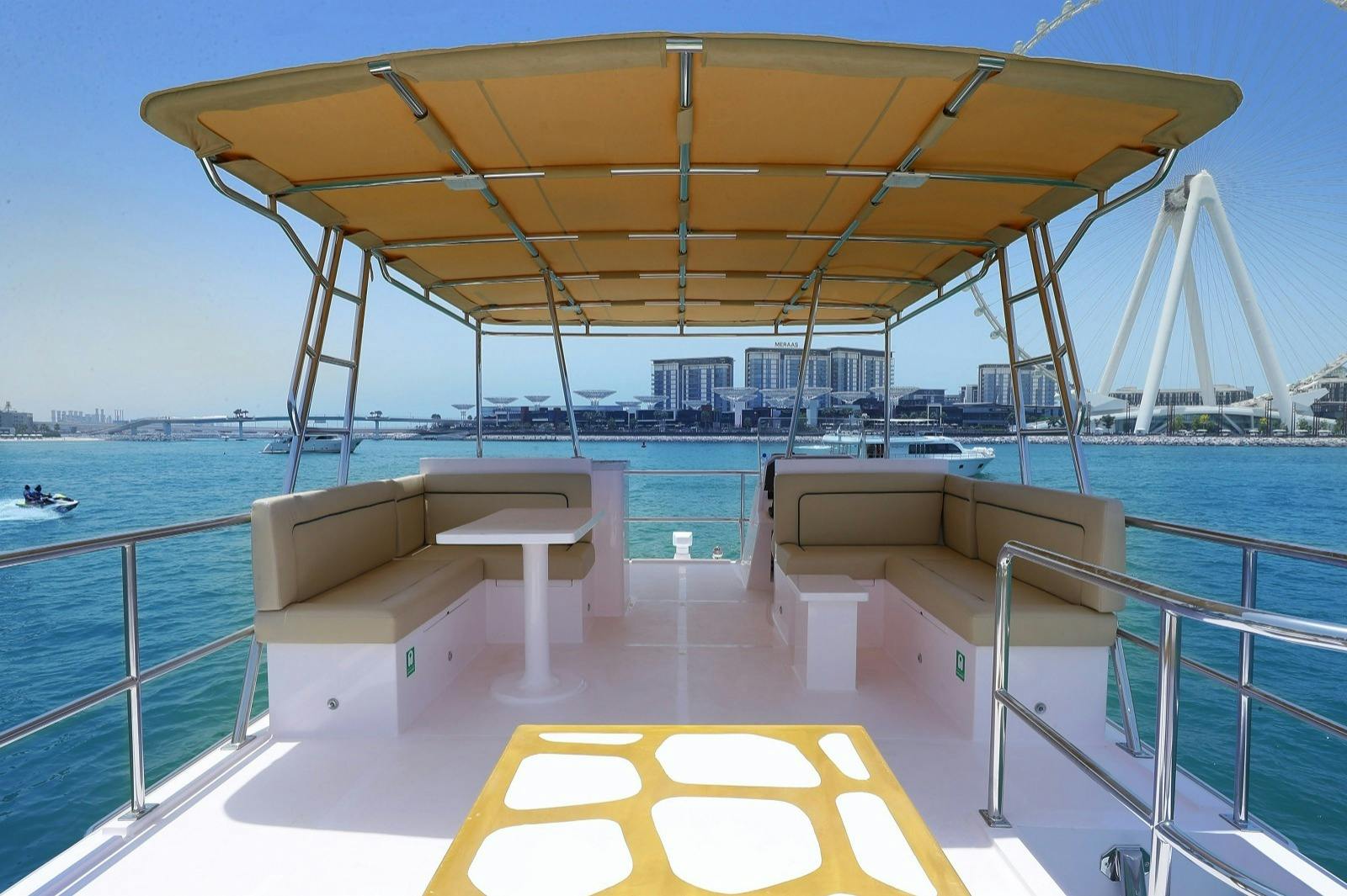 A boat's upper deck with a shaded seating area, featuring beige benches and a central table, overlooking a coastal cityscape.