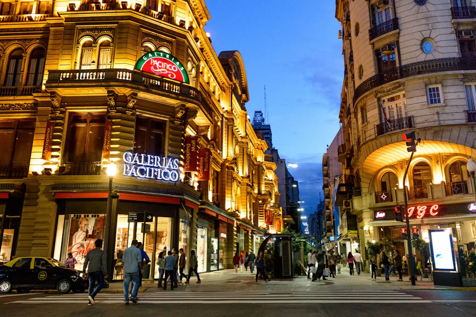 A busy pedestrian street at dusk with illuminated buildings, including Galerías Pacífico, and people walking.
