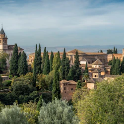A scenic view of a historic complex with tall cypress trees, rustic buildings, and a distant mountain range under a partly cloudy sky.