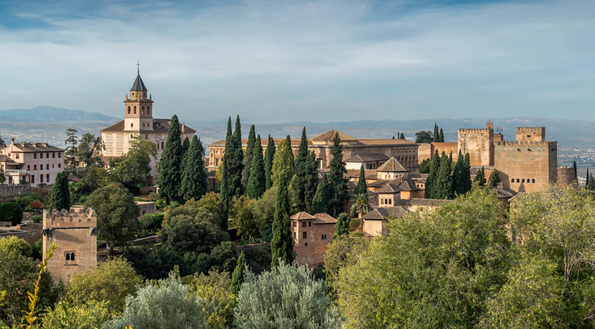 A scenic view of a historic complex with tall cypress trees, rustic buildings, and a distant mountain range under a partly cloudy sky.
