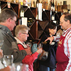 En man i rött förkläde häller vin för flera personer i en vinbutik med hyllor av vinflaskor i bakgrunden.