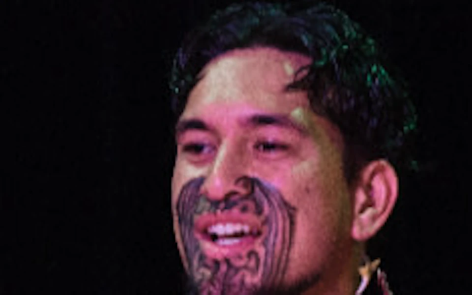 A man in traditional attire plays an acoustic guitar on stage, flanked by others in similar outfits, against a dark background.