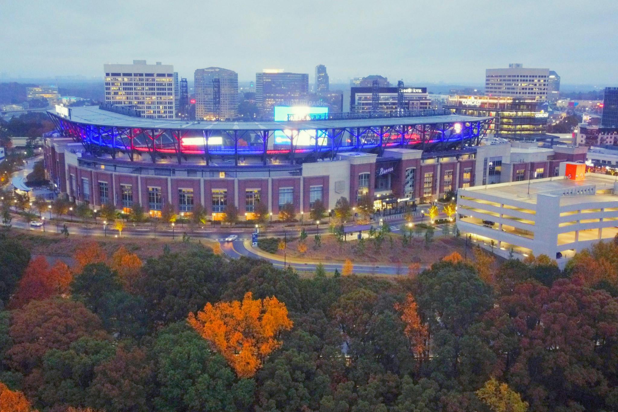 Вид с воздуха на ярко освещенный стадион с разноцветными огнями, окруженный деревьями с осенней листвой и городскими зданиями на заднем плане.
