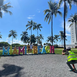 People posing in front of colorful "Miami Beach" sign under palm trees, with a photographer capturing the moment.
