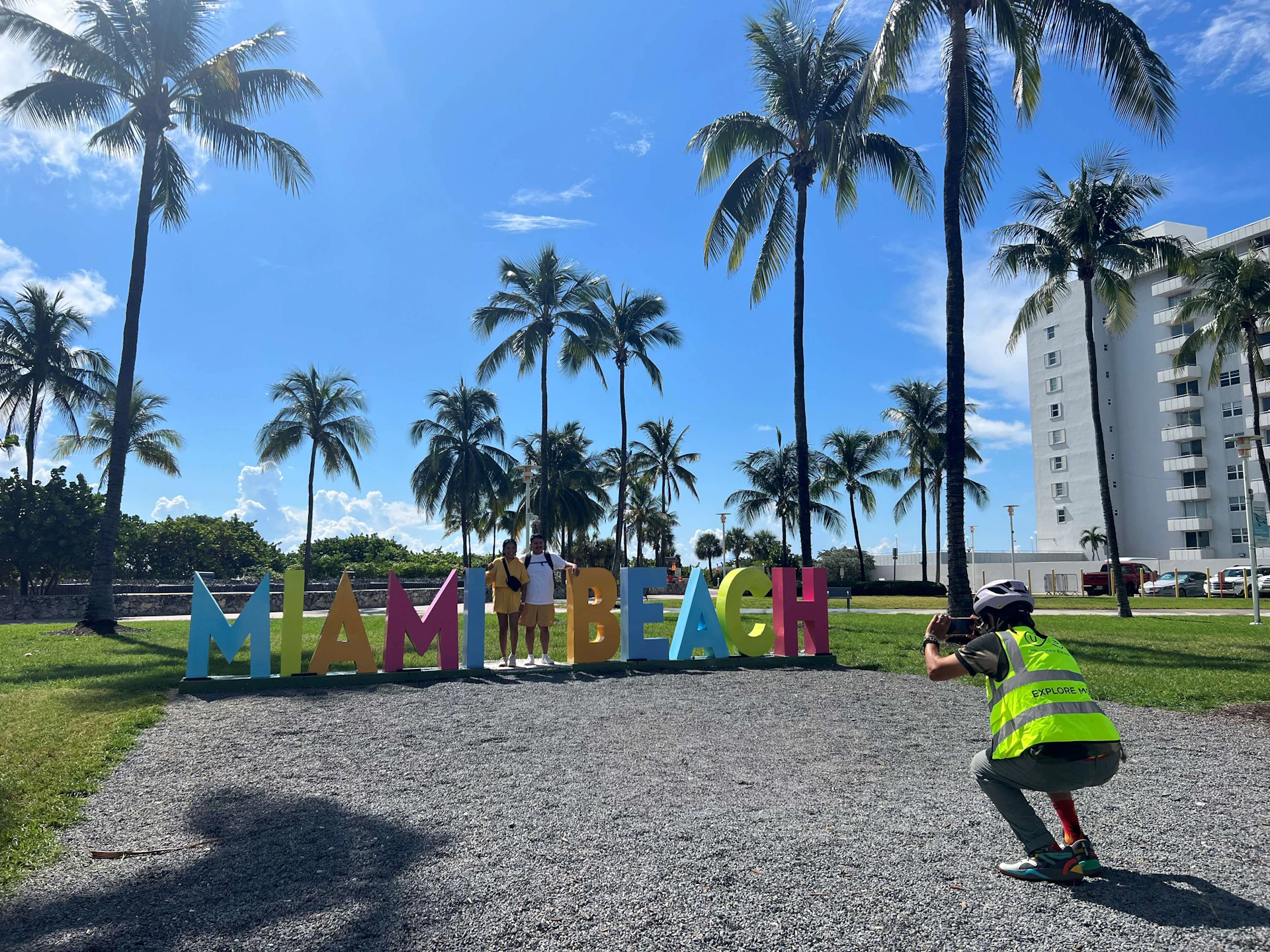 People posing in front of colorful "Miami Beach" sign under palm trees, with a photographer capturing the moment.