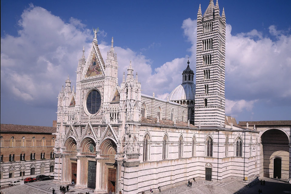 A Gothic cathedral with a striped facade, rose window, and tall bell tower under a partly cloudy sky.