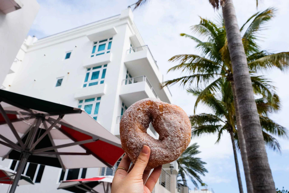 A hand holds a powdered donut in front of a white multi-story building with blue windows and palm trees.