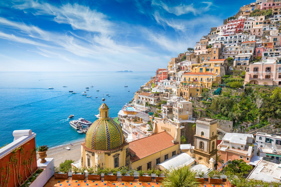 Colorful hillside buildings overlook a serene coastline with boats floating on a bright blue sea under a clear sky.