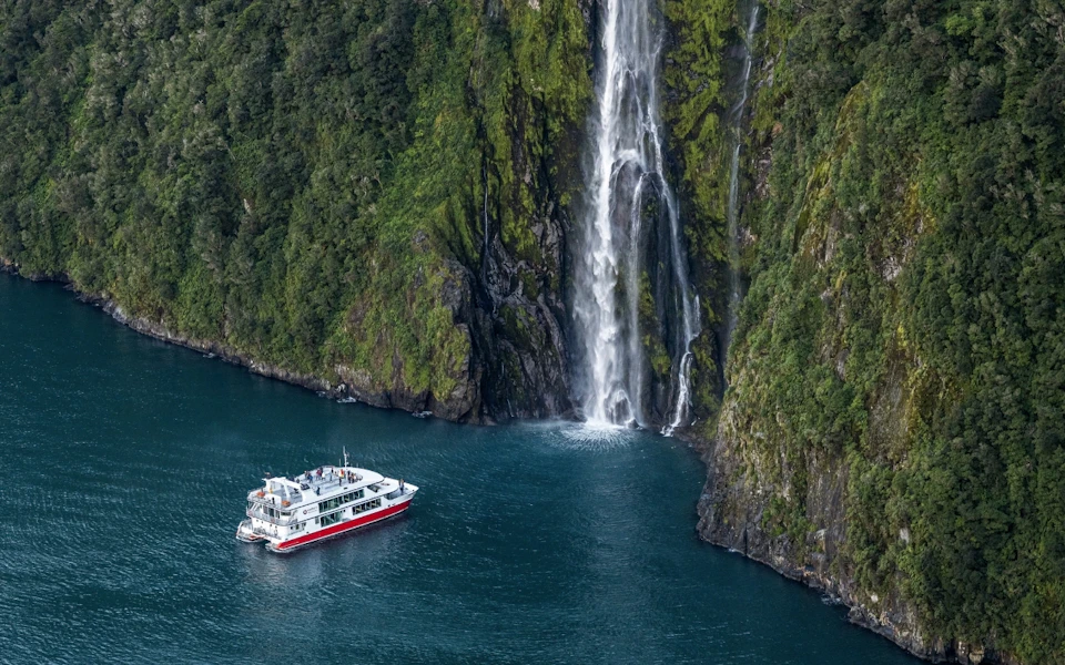 A boat floating near a tall waterfall cascading down a lush green cliff into a calm body of water.