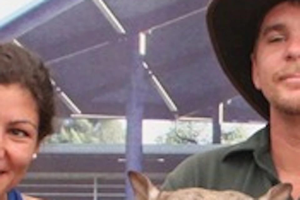 Two people sitting on a bench in front of a sign for Caversham Wildlife Park, holding a wombat between them.