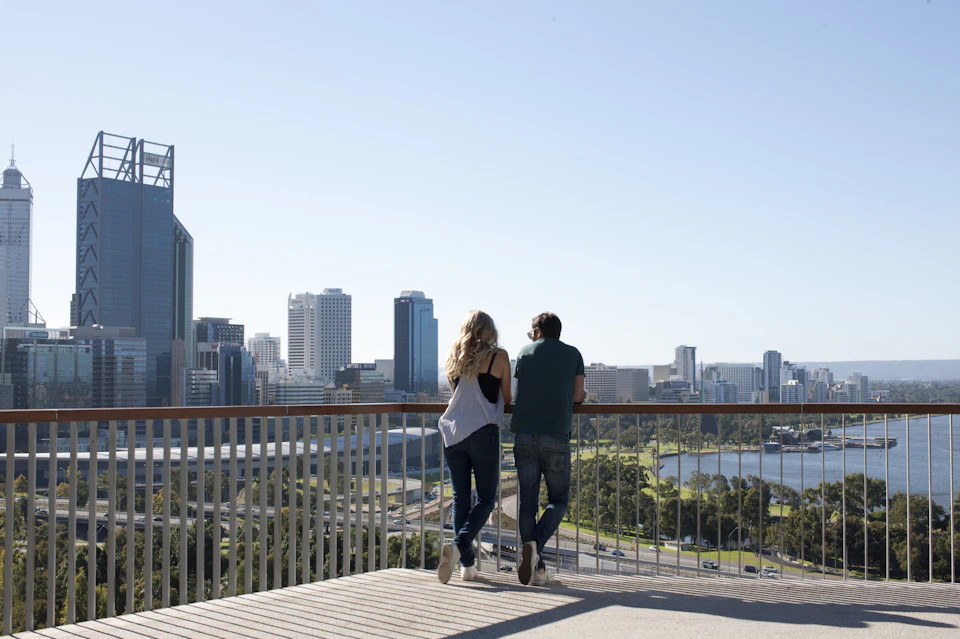 Two people leaning on a railing, overlooking a cityscape with tall buildings and a river under a clear sky.