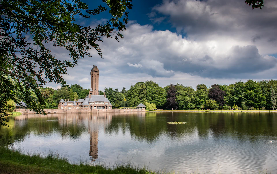 A tall, beige tower and building are reflected in a calm lake, surrounded by lush greenery under a partly cloudy sky.