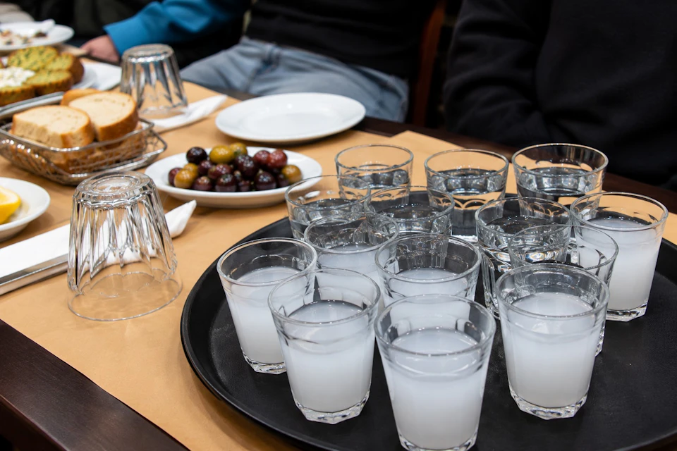 A table with a tray of glasses filled with a clear beverage, a plate of olives, bread, and plates in a casual dining setting.