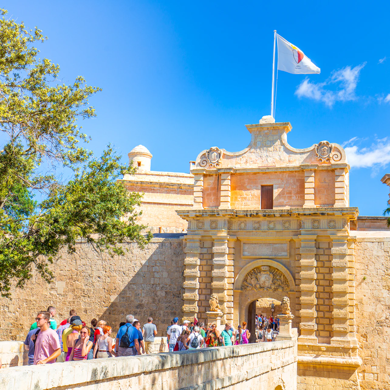 Tourists walk through the entrance gate of an ancient stone fortress under a clear blue sky, with a flag flying atop the gate.