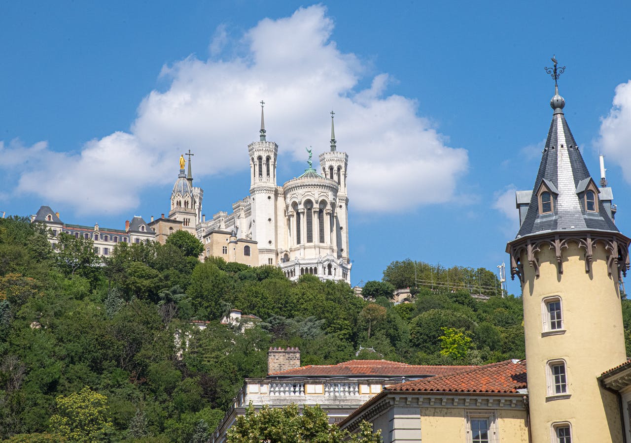 En vit basilika med tvillingtorn som ligger på en kulle, omgiven av grönska, under en blå himmel med moln.