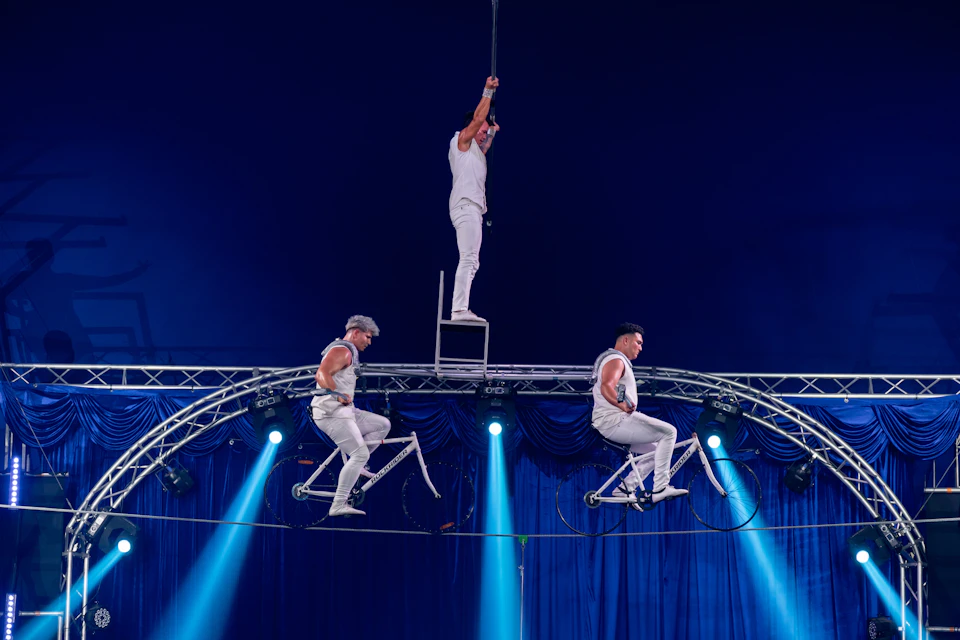 Three acrobats in white perform on a tightrope. Two ride bicycles while one balances on a chair, under stage lighting.