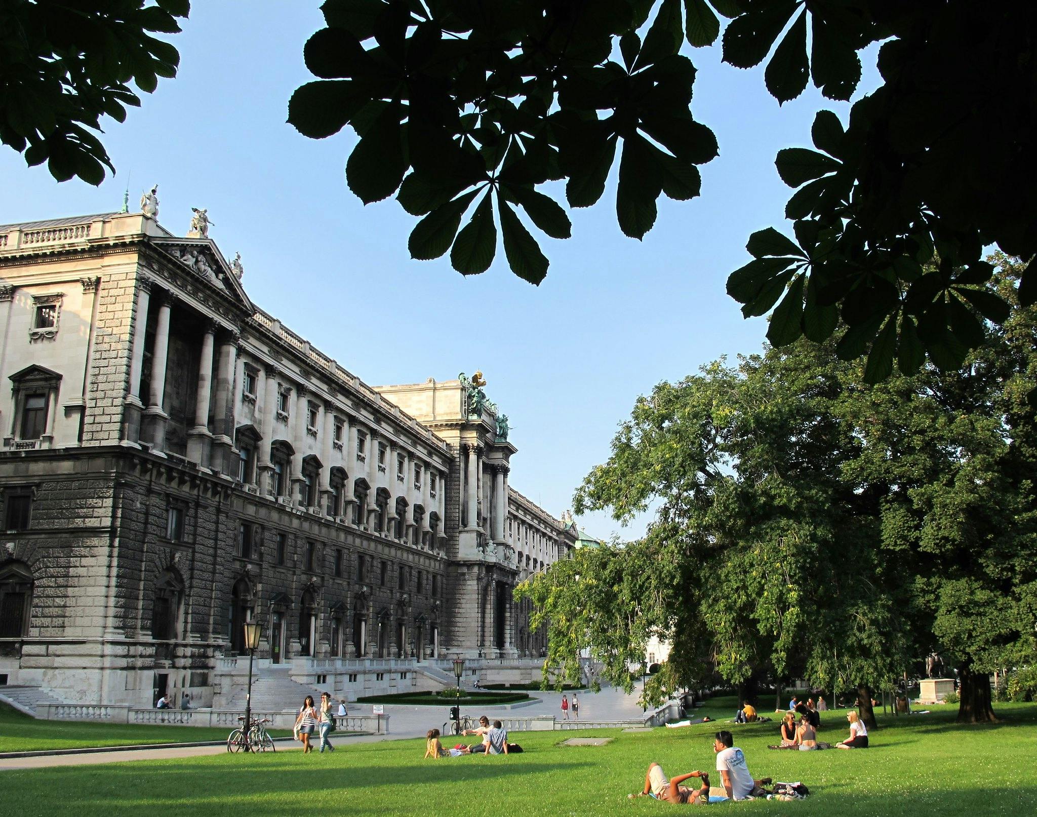 Folk slappnar av på gräset framför en storslagen, historisk byggnad med pelare och statyer, inramad av träd och en klarblå himmel.