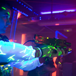 Person i ett mörkt rum som håller en laserpistol, upplyst av neon och svart ljus med glödande mönster på sina kläder.