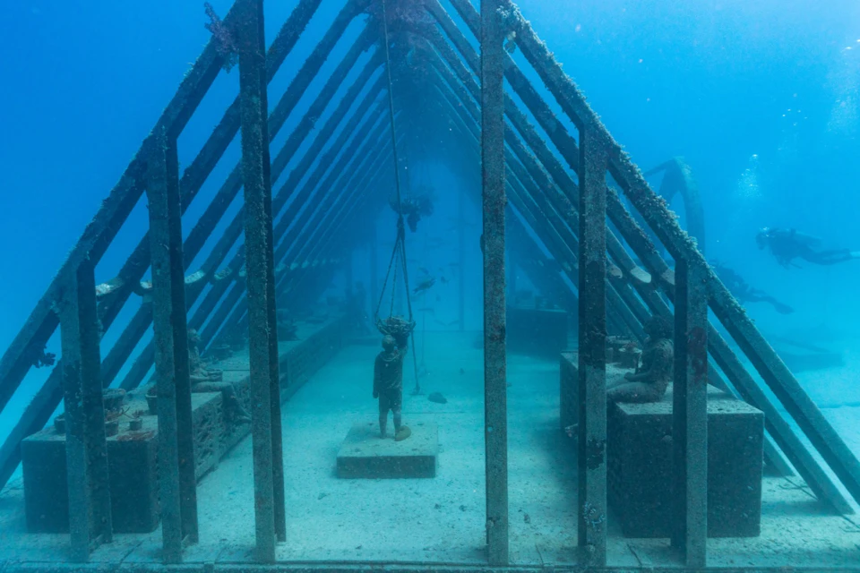 Underwater structure resembling a chapel with a statue of a figure under a metal framework, surrounded by marine life.