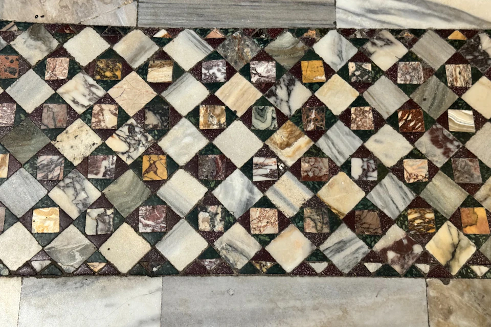 A mosaic floor pattern composed of small square tiles in various colors, including beige, brown, green, and marbled textures.