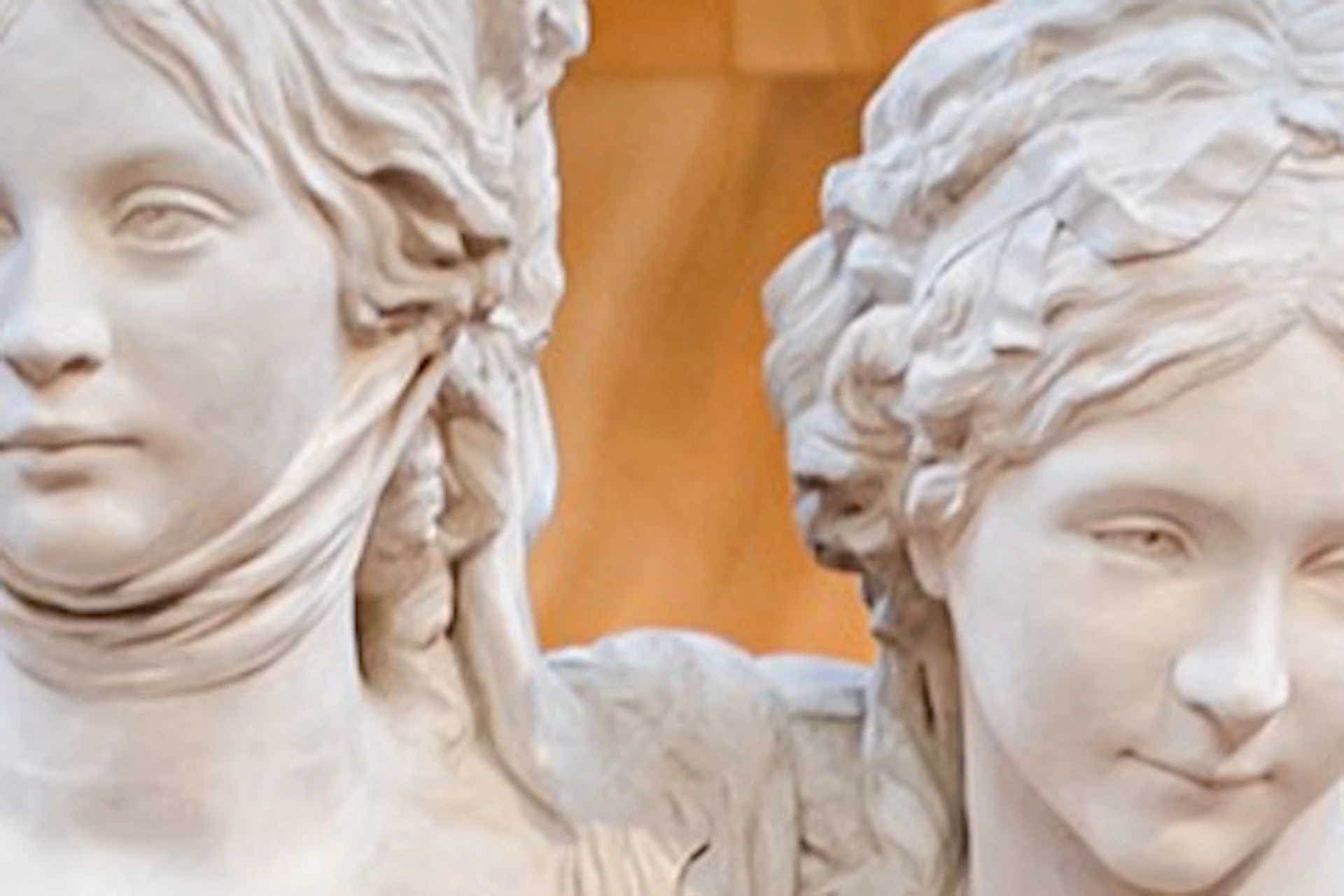 A marble statue of two women standing together in a museum, with other sculptures visible in the background.