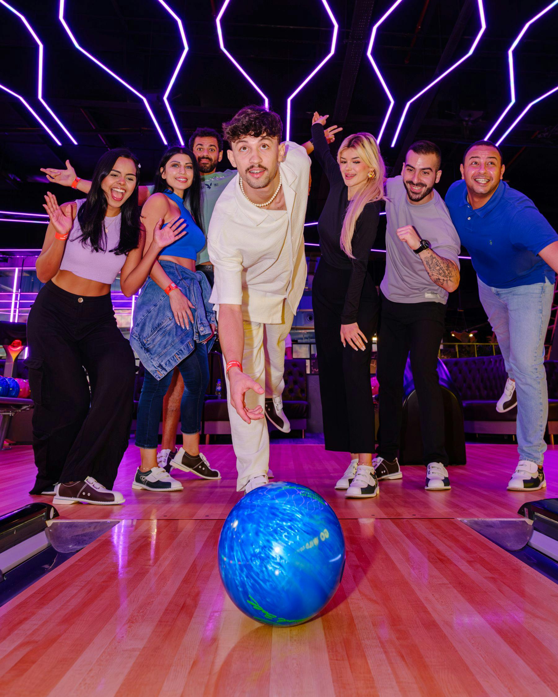 Μια ομάδα ανθρώπων σε μια αίθουσα μπόουλινγκ, ένα άτομο που παίζει μπόουλινγκ με μια μπλε μπάλα, φώτα νέον ορατά στο παρασκήνιο.